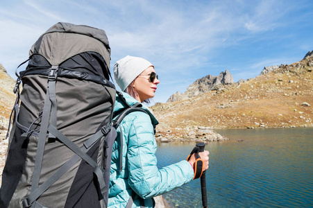 太阳镜和帽子与跟踪在她手中的徒步背包和山齿轮的徒步旅行者女孩看着美丽的群岛，北高加索高山湖泊风景