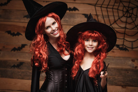 万圣节的概念   美丽的白人母亲和她的女儿在女巫服饰，魔杖庆祝万圣节假扮与蝙蝠和蜘蛛网木制演播室背景上的长长的红头发