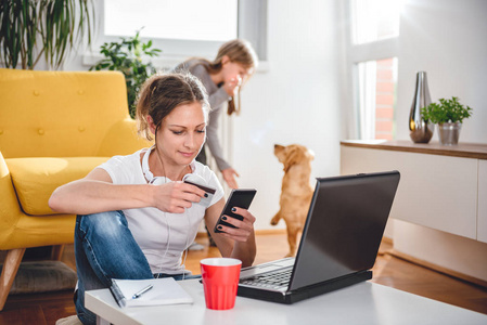 女人穿着白色衬衫和耳机，在智能手机上网上购物，并在后台与女儿和狗使用信用卡。