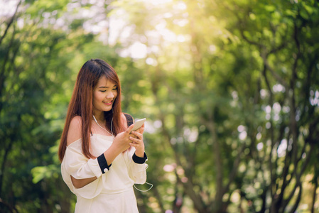 可爱的亚裔女子在公园里坐在温暖的春日，华丽女听音乐的耳机和细胞电话信息检索阅读愉快的短信在手机上