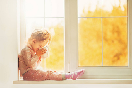 可爱的孩子的小姑娘在睡衣坐在大窗口玩微笑回家享受。可可杯站在窗口上。秋季赛季室内家庭纺织行业节日人们观念