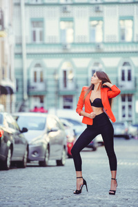 时尚的外观, 炎热的一天模型的一个年轻女子走在城市, 身穿红色夹克和黑色的裤子, 金发碧眼的户外城市温暖的背景