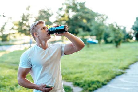 一个男人在锻炼时 atlen 喝水, 手里拿着耳机, 手持智能手机。在这个城市的夏天, 一种生活方式。蛋白振动筛