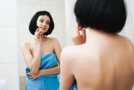 美丽迷人的女孩与短的黑发在一条蓝色毛巾看自己在镜子