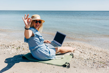 年轻女子穿着蓝色连衣裙，坐在海滩上，怀里抱着笔记本电脑，背景是大海