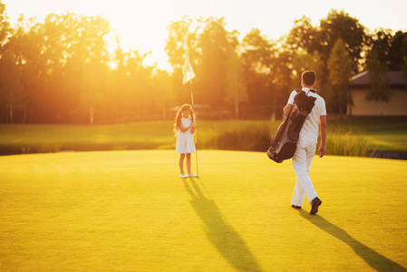 一个穿白色西装的男人和一个高尔夫球棍包向在日落时站在洞口旁边的女孩走去。