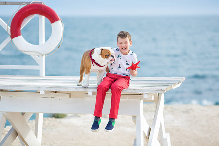 英俊的男孩青少年 happyly 花时间与他的朋友斗牛犬在海边的孩子狗控股玩两个海星接近生活浮标穿红色长裤拖鞋和 tshirt