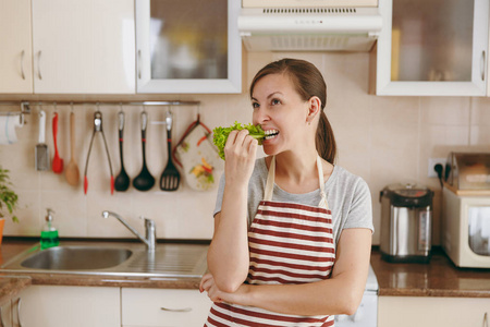 一个年轻漂亮的女人在她的嘴在厨房的围裙与莴苣叶。节食概念。健康的生活方式。在家做饭。准备食物