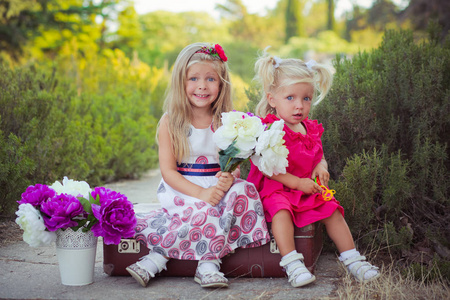 姐妹两个金发碧眼深冰蓝眼睛穿着五颜六色的礼服摆在中央公园森林草甸坐在旅行树干袋与花束紫罗兰紫色花