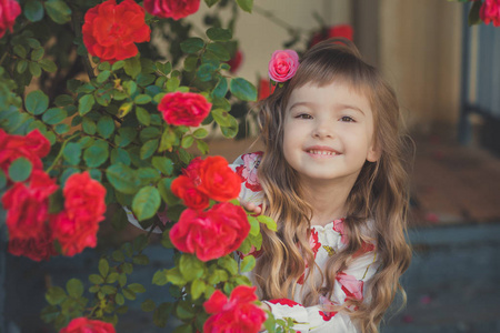 可爱的女婴与金发碧眼的卷发和幸福的明亮的儿童眼睛摆在中央公园靠近巨大的灌木花红色白色粉红色的玫瑰穿夏天时髦的衣服