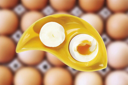 煮鸡蛋在一个黄色的蛋杯中叠加在一盒棕色的鸡蛋上，构成射击