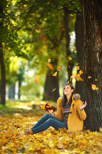 在一个温暖的日子里, 在秋天的城市公园里, 一个穿着黄色外套和牛仔裤坐在枫树下, 带着一本红色的书的美丽快乐微笑的棕色头发的女人