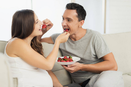 一个孕妇躺在轻便的沙发上。一个男人坐在她旁边。他们吃草莓和甜食