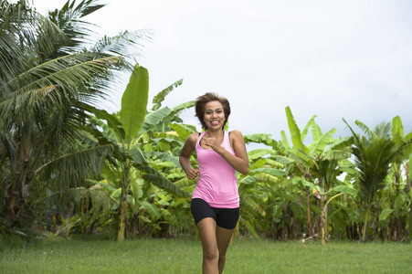 年轻有吸引力的亚洲体育赛跑者妇女奔跑在密林微笑的愉快训练在草本与棕榈树的背景的锻炼