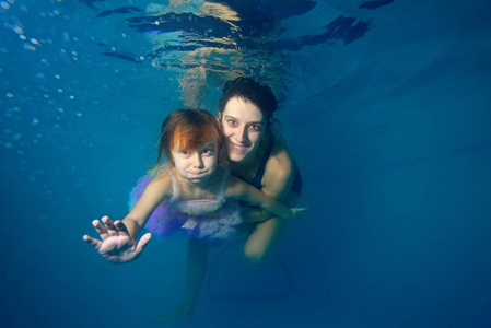 小女孩和指导员在水下游泳，微笑着，在蓝色的背景下看着相机。肖像。特写镜头。从水下看到的景色。景观导向