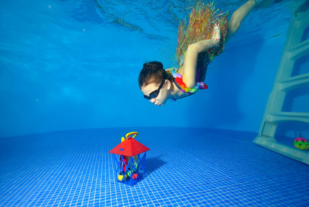 穿着狂欢节服装的运动小女孩潜入池底，获得玩具礼物。肖像。在水下射击。景观导向
