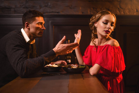 年轻夫妇在餐馆的争吵，糟糕的浪漫约会。 穿着红衣服的优雅女人和英俊的男人一起吃饭