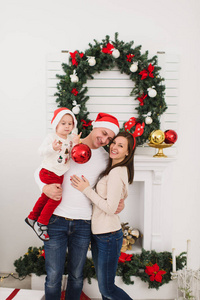 快乐的父母带着可爱的小儿子戴帽子。在圣诞花环的背景下, 孩子带着大红树玩具球站在装饰一新的新年灯房里。家庭, 假日2018概念