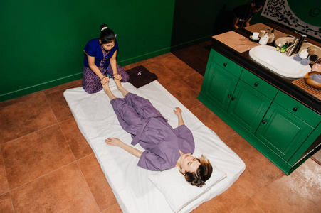 年轻的泰国男按摩师女孩在东方异国情调的礼服制作和演示不同的传统 spa 仪式的程序欧洲美女穿着睡衣在绿色放松瑜伽工作室