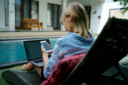 从后面的照片上可以看到一位金发女性在暑假期间坐在一座豪华的热带别墅里，坐在游泳池旁打开的便携式电脑上看着手机显示器。