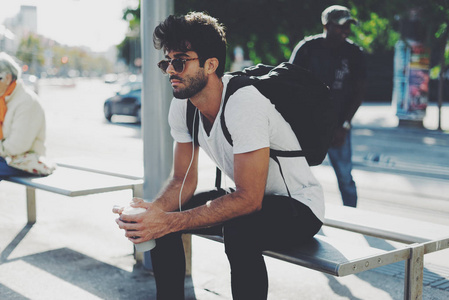 年轻的胡须男，卷发，黑发，戴着太阳镜，正在车站等火车。 时髦的家伙坐在长凳上，背上背着一个背包，背景模糊。