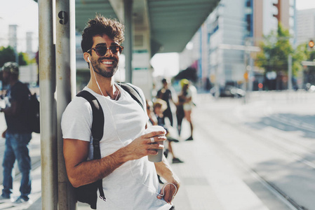 英俊的男性，戴着太阳镜，带着深色卷发，在阳光明媚的日子里，站在街上喝杯咖啡时，正对着相机微笑。 胡须男在车站等交通工具。