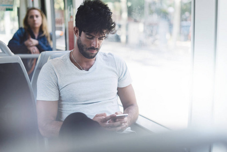 留胡子的嬉皮士正在一台智能手机的显示器上键入电子邮件，该智能手机连接到公共Wi Fi，而坐在城市公共汽车上。 英俊的男性正在看手