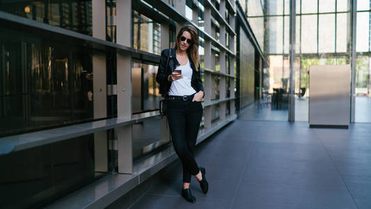 迷人的金发女郎穿着黑色牛仔裤皮夹克和太阳镜，站在办公室入口区的玻璃和钢墙旁看着智能手机显示器。