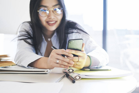 裁剪照片，重点是一个手机在一个美丽的亚洲女性手中戴着眼镜的背景。 年轻的商务女性正在智能手机屏幕上阅读信息。