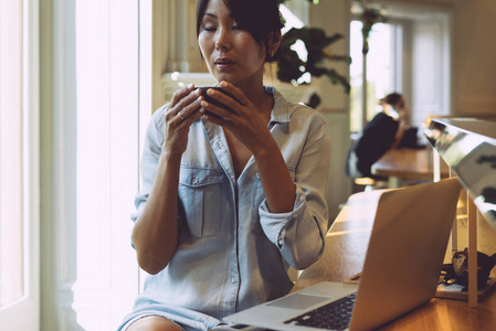 美丽的亚洲女人穿着浅蓝色牛仔裤连衣裙，一边喝茶，一边坐在桌子上，桌上放着一台便携式电脑。 年轻的自由职业者正在一个现代的合作空间
