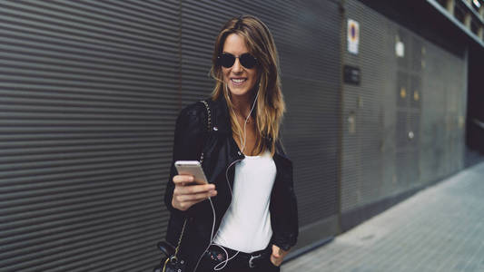 开朗的年轻女人戴着太阳镜微笑，一边看朋友的留言。 时尚的女性在线聊天，而站在灰色的墙壁背景上的手机在一条街上。