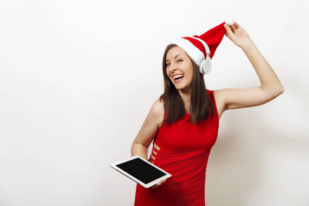 漂亮的年轻快乐的女人穿着红色的裙子, 戴着圣诞帽, 戴着耳机听音乐, 在白色背景上拿着平板电脑。孤立的圣诞老人女孩小工具。新年假