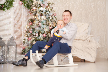 充满爱心的父亲和年幼的小儿子在一棵圣诞树旁的白色木制雪橇上玩耍，上面有新年礼物盒