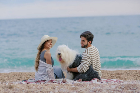一位戴草帽的年轻女子和一个蓄着胡须的男人在海上玩着一只毛茸茸的狗，坐在格子呢上
