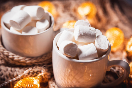 圣诞节背景与可可粉或咖啡与棉花糖在一个白色杯子在棕色针织冬天围巾和发光的金黄花环。家居舒适温馨的美丽理念
