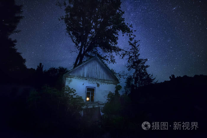古老的破旧的房子在森林中,在壮丽的星空和坠落的星星下,窗户里有光.