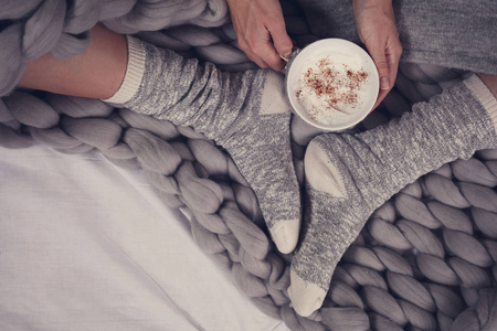 女性脚穿舒适保暖的羊毛袜子接近。裹着温暖毛毯的女人在床上喝咖啡