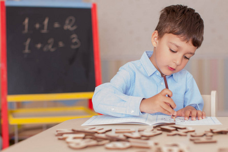 可爱的小男孩穿着蓝色衬衫坐在桌子旁学习。木制的字母，数字，书的拷贝本和桌子上的彩色钢笔和铅笔..模糊的墙壁和黑板背景