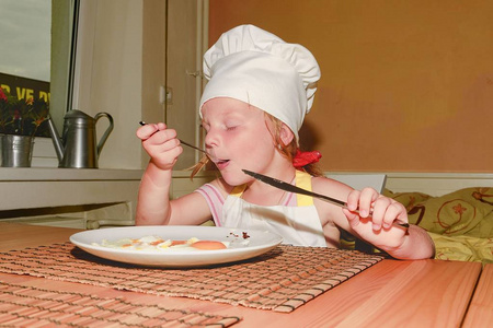 小女孩有点心。小女孩吃火腿和鸡蛋。可爱的女孩穿着白色的厨师服装。家庭和童年概念