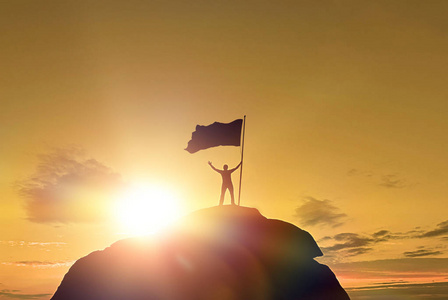 高成就轮廓的男子胜利旗帜在山顶上举起双手。 山顶上的人。 概念设计。 在日落时乌云密布的戏剧性天空上。