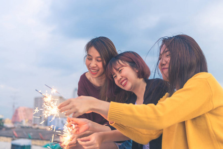 户外拍摄的年轻人在屋顶的聚会。快乐的亚洲女朋友们在傍晚的日落时分享受和玩烟火的屋顶派对。节日庆典节日聚会。青少年生活方式党