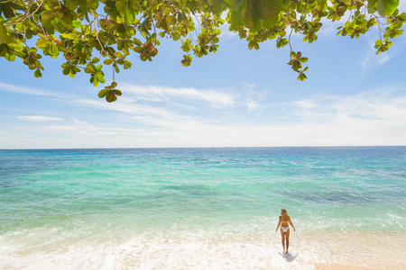 穿白色泳装的女孩站在海滩上。 背景上的蓝天和绿叶。