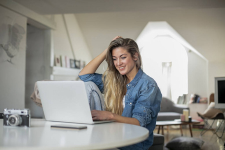 年轻漂亮的女人在家里为一台电脑工作，她的笔记本电脑放在白色的桌子上，做自由职业者