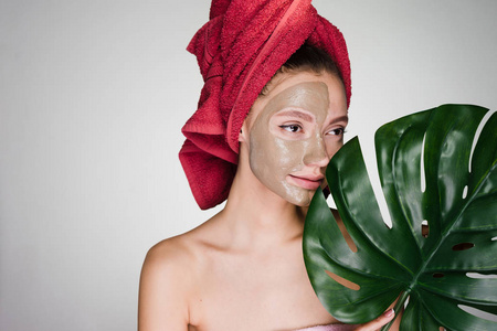 年轻美丽的女孩, 她的头上有一条红色的毛巾, 在治疗粘土的半边脸上, 保持一片绿叶