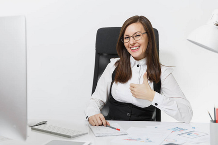 美丽的微笑棕色头发业务妇女在西装和眼镜坐在办公桌上, 在计算机与现代显示器与文件在轻型办公室, 显示拇指在白色背景