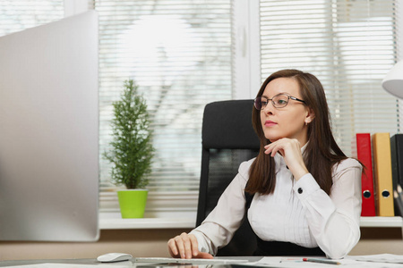 美丽的严重和全神贯注的棕色头发的业务妇女在西装和眼镜坐在办公桌与片剂, 工作在计算机与现代显示器与文件在轻型办公室, 看一边