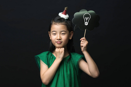 可爱的亚洲女孩穿着一件绿色连衣裙，手里拿着一根创意灯泡棒，黑色背景上有一个圣诞头装饰。 思考的表达