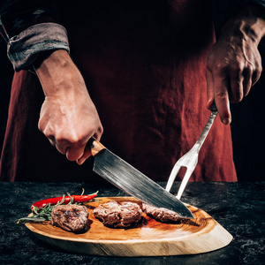 在木板上用肉叉和刀切成美味的烤牛排，配上迷迭香和辣椒