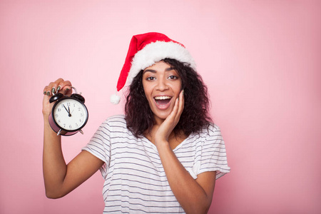 快乐的年轻女子在圣诞老人的帽子举行时钟。混合种族非洲妇女在粉红色的圣诞