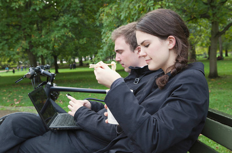 迷人的高加索年轻夫妇坐在公园的长凳上。男人用笔记本电脑，女人吃面条